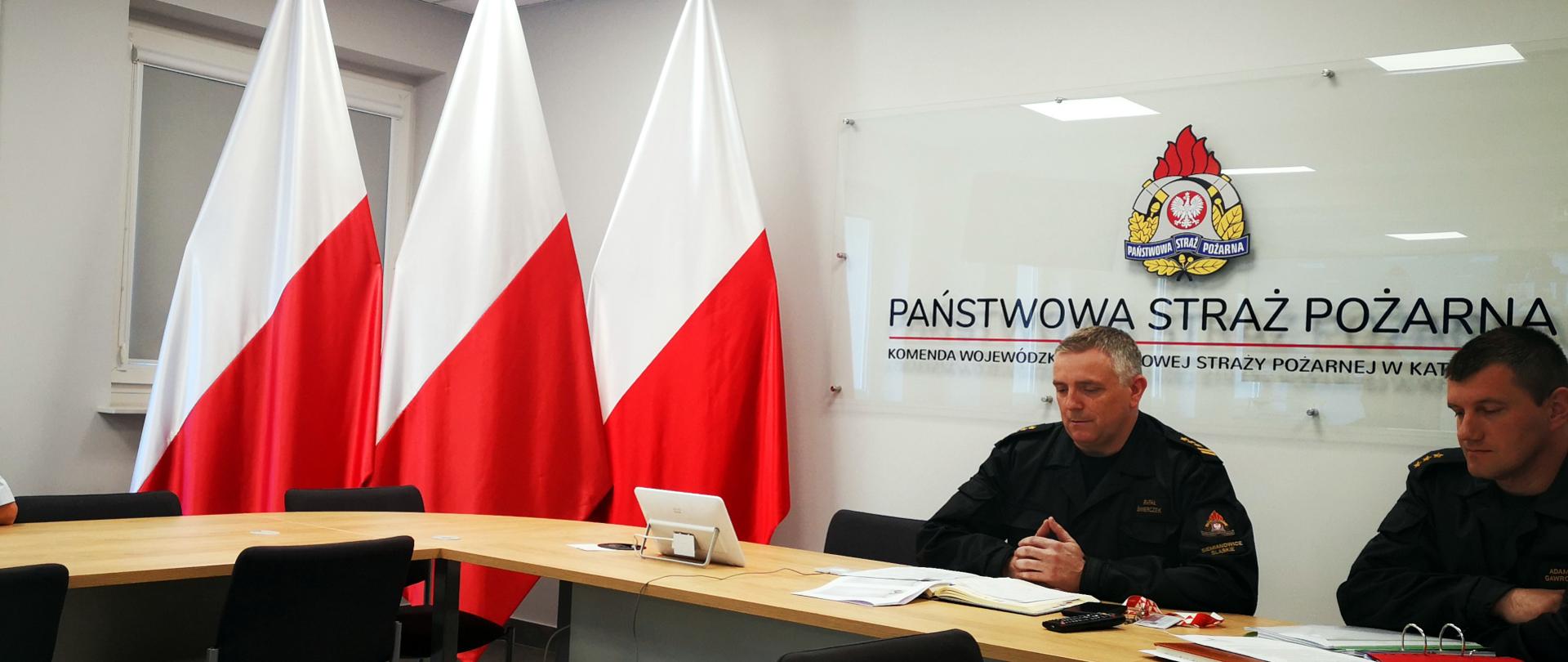 Na zdjęciu sala sztabowa. Zastępca Śląskiego Komendanta Wojewódzkiego PSP st. bryg. Rafał Świerczek prowadzi wideokonferencję z kadrą kierowniczą PSP woj. śląskiego. Po prawej stronie siedzi Naczelnik Ośrodka Szkolenia Komendy Wojewódzkiej. W tle po lewej stronie ustawione trzy flagi narodowe. Na ścianie napis Państwowa Straż Pożarna i logo PSP.