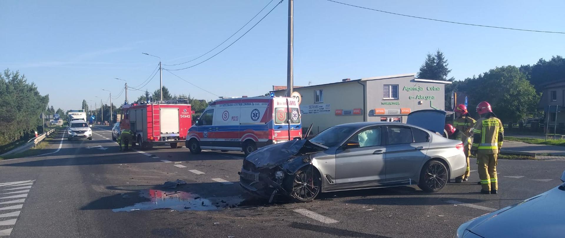 Zdjęcie przedstawia kolizję drogową na DK 11 z udziałem 2 samochodów osobowych. Na zdjęciu po prawej stronie widać uszkodzony samochód osobowy po kolizji natomiast na drugim planie widzimy pojazd ratowniczo z JRG Ostrzeszów i tuż za nim karetkę pogotowia ratunkowego. Zdjęcie wykonane w porze dziennej na drodze.