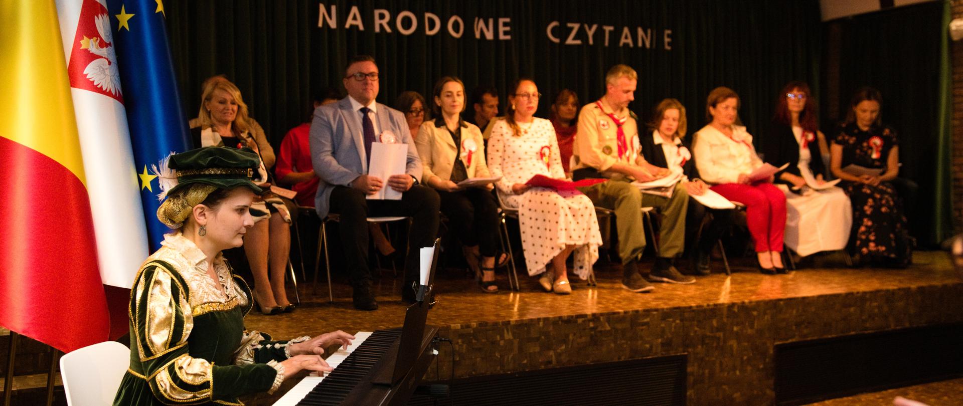 The National Reading speakers sit on stage. In the foreground, a woman in period costume accompanies the reading on an electric piano.
