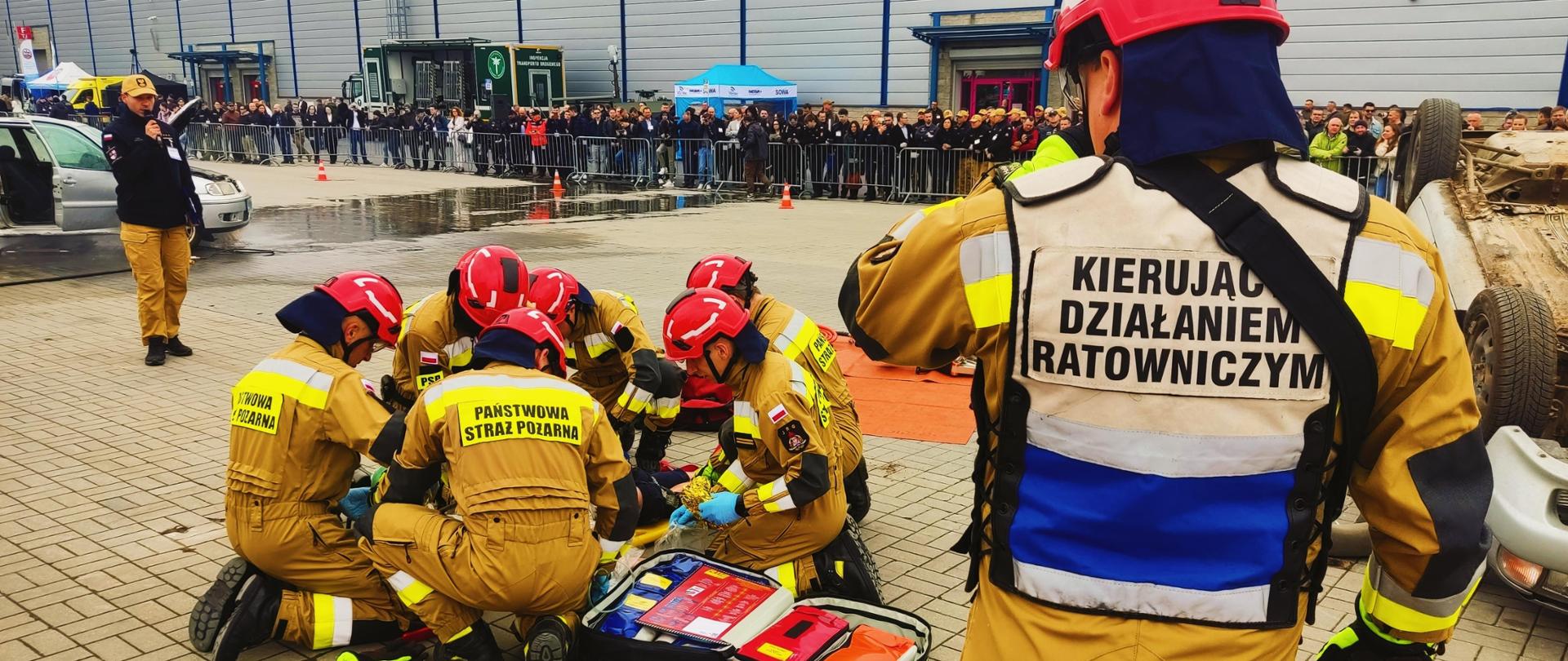 Na zdjęciu widzimy strażaków podczas ćwiczeń z ratownictwa technicznego. W tle widzimy widzów. 