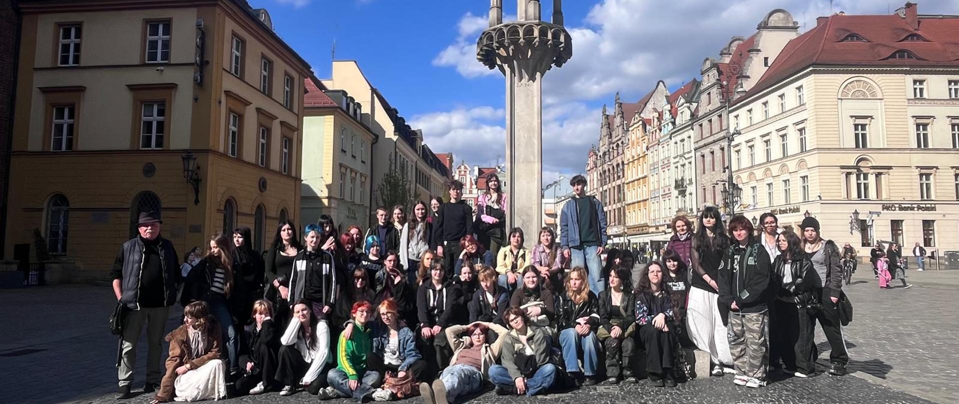Zdjęcie jest pamiątkową fotografią grupową wykonaną podczas wycieczki do Wrocławia. Uczestnicy stoją na rynku pod pręgieżem.