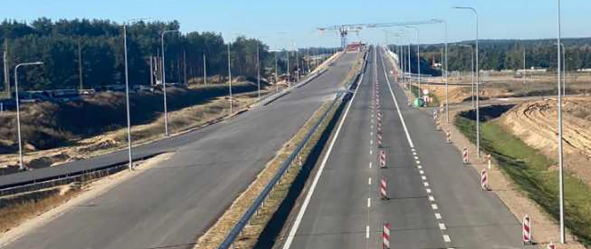 Droga ekspresowa z pachołkami i dźwigiem w tle, otoczona zielenią i oświetleniem ulicznym.