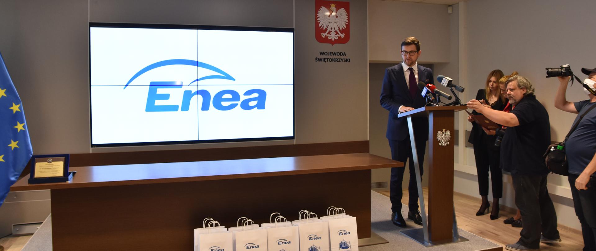 Minister Andrzej Śliwka stoi przy mównicy, po lewej stronie ekran z logo Enei, po prawej stojący obok ministra dziennikarze