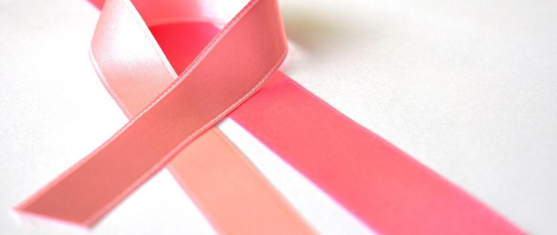 Na zdjęciu znajduje się różowa wstążka, która jest spleciona w charakterystyczny sposób dla akcji propagującej miesiąc świadomości raka piersi. Wstążka znajduje się na białej powierzchni.