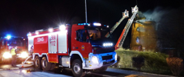 Na pierwszym planie na chodniku stoi samochód pożarniczy Ochotniczej Straży Pożarnej w Nowem do którego podłączone są węże, ma wysunięty masz oświetleniowy i oświetla teren za nim gdzie w tle widać dwa podnośniki strażackie z których strażacy dogaszają pożar wierzy wiatraka. Za samochodem widać przód kolejnego pojazdu straży pożarnej.