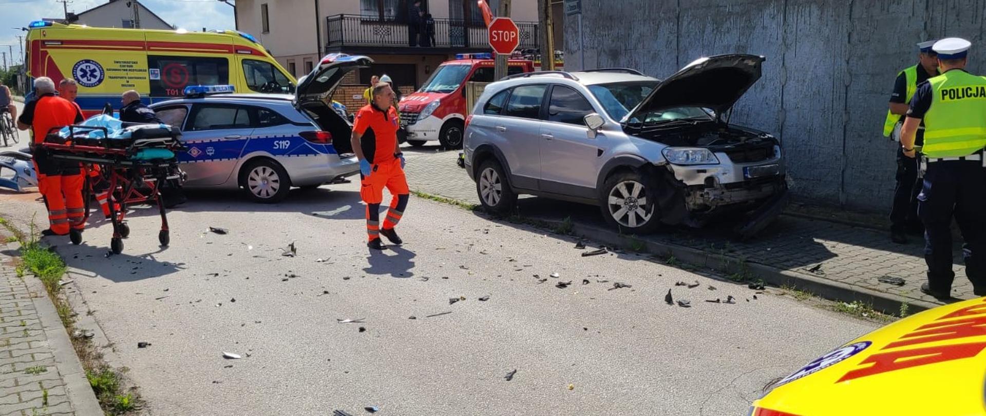 Zdjęcie przedstawia miejsce wypadku: z lewej strony nosze na tle zniszczonego radiowozu, za nim ambulans. Z lewej strony zniszczony samochód osobowy, a za nim samochód strażacki. Przez drogę przechodzi ratownik medyczny w stronę dwóch policjantów.