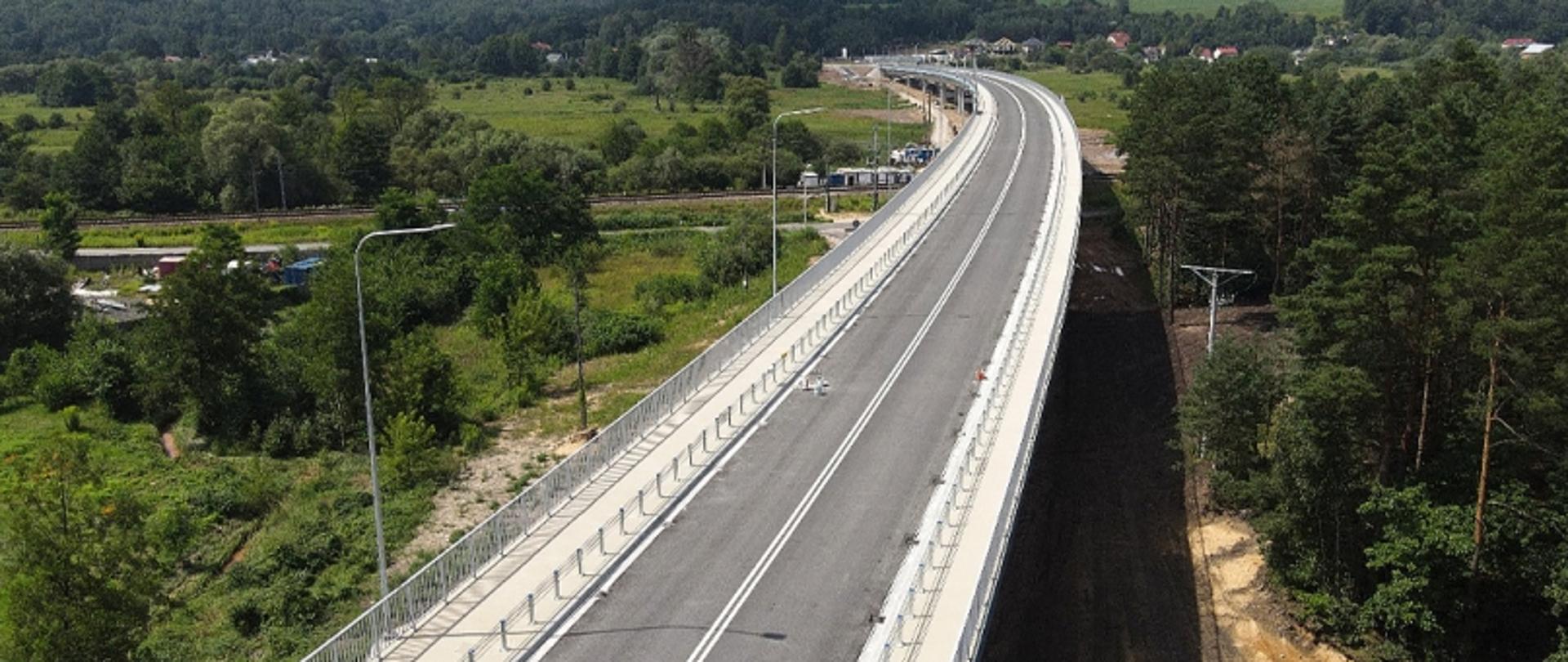 Estakada z czterema pasami ruchu, latarniami i barierami, otoczona lasem i budynkami w oddali.