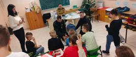 w klasie przy biurku siedzi strażak dwaj strażacy klęczą na podłodze przy nich dziecko kilka dzieci siedzi przed nimi na krzesełka