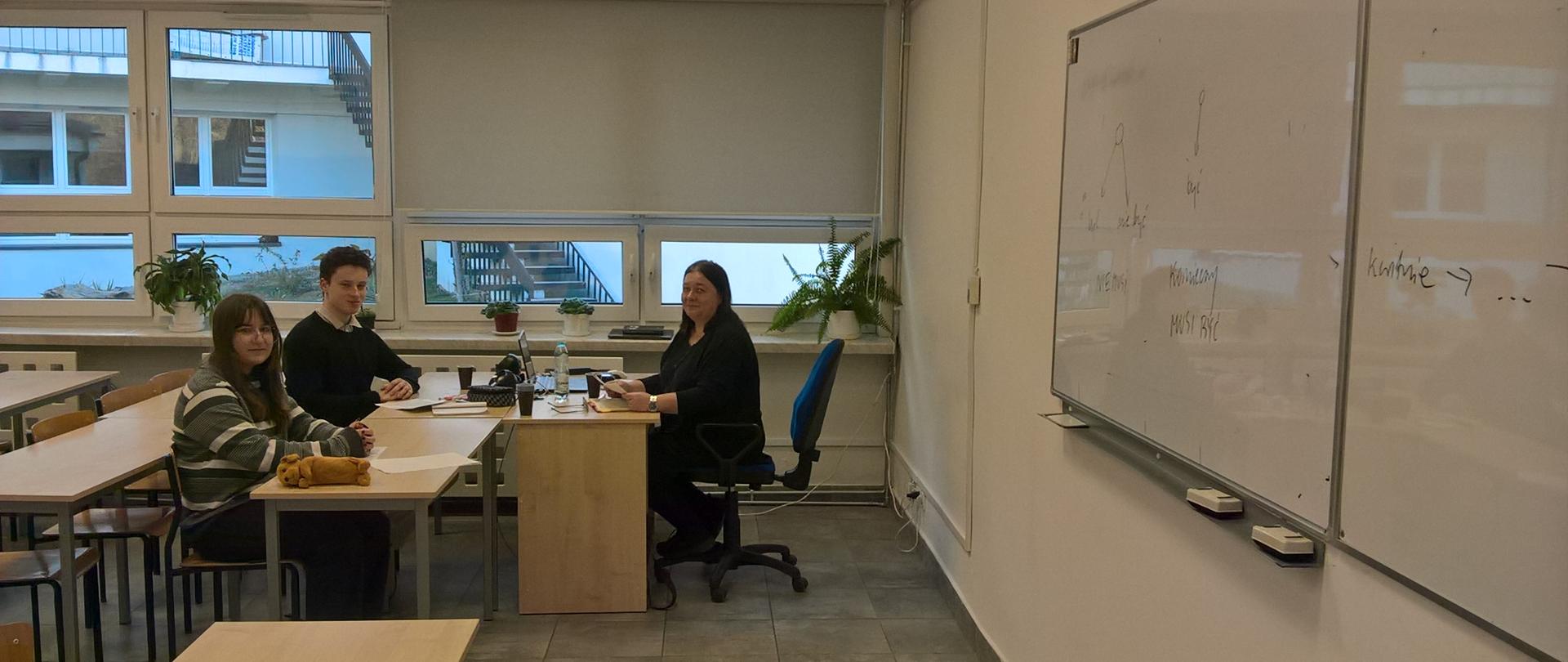 W sali lekcyjnej PLSP w Lublinie. Anna Palusińska siedzi za biurkiem i prowadzi wykład. Na przeciwko siedzą w ławkach uczeń i uczennica.