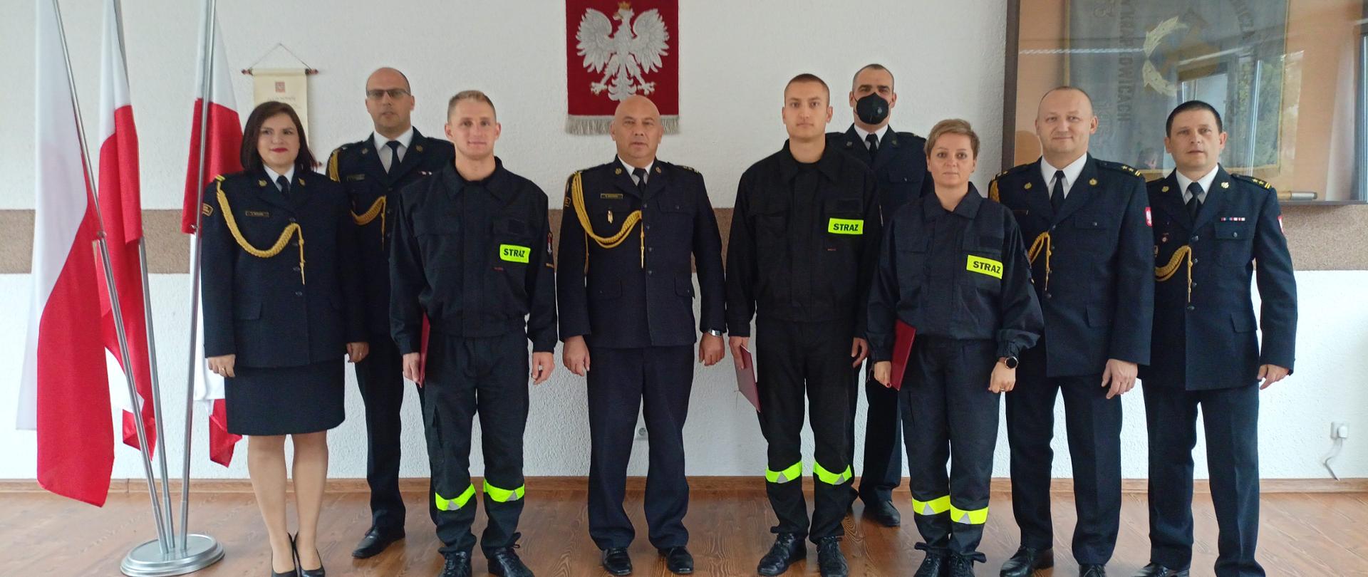 Zdjęcie grupowe, na którym stają funkcjonariusze składający ślubowanie oraz kadra KP PSP w Krapkowicach. W tle na ścianie Godło RP, po lewej stojak z flagami narodowymi RP. Kadra komendy w umundurowaniu galowym, strażacy w koszarowym. 