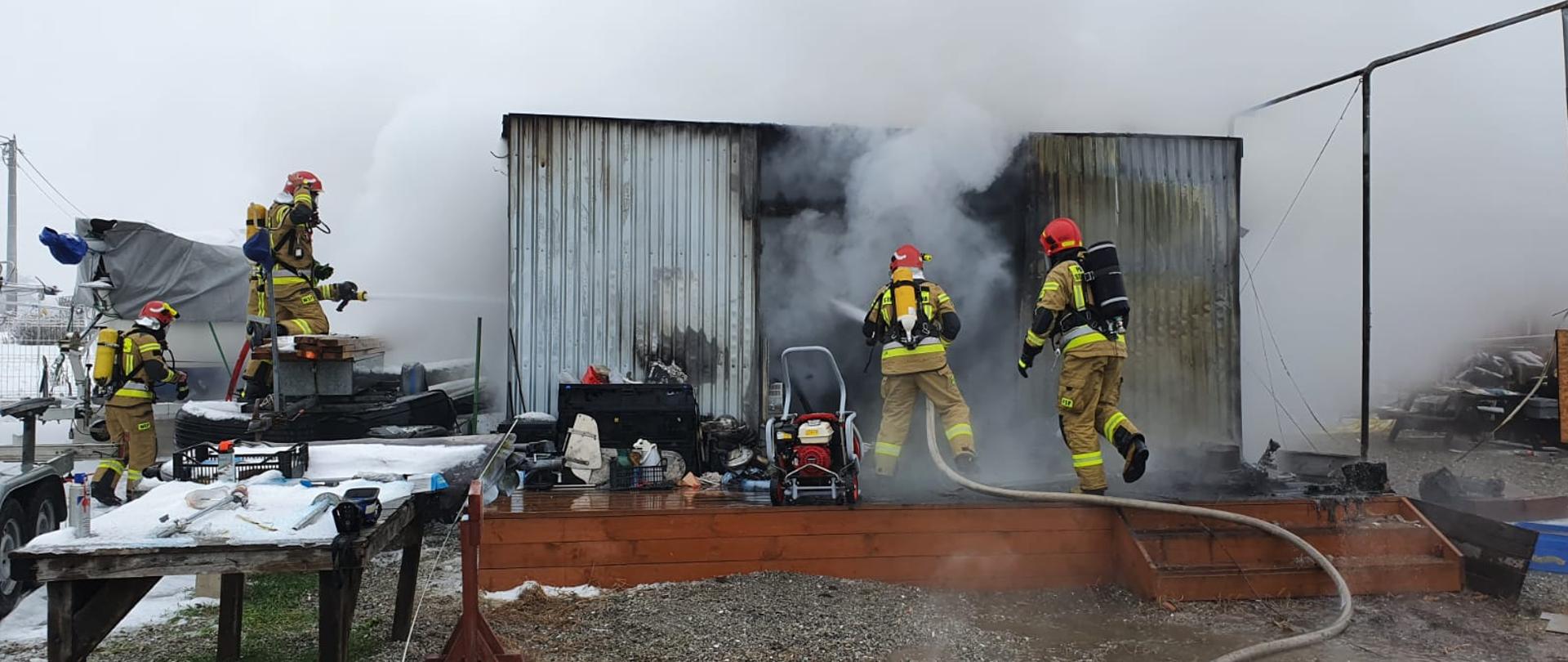 Blaszany budynek gospodarczy czterech strażaków w ubraniach specjalnych oraz sprzęcie ODO gaszą pożar budynku przed nim na brązowym podeście ustawiony czerwony wentylator wokół budynku różnego rodzaju sprzęt widoczny biały dym.