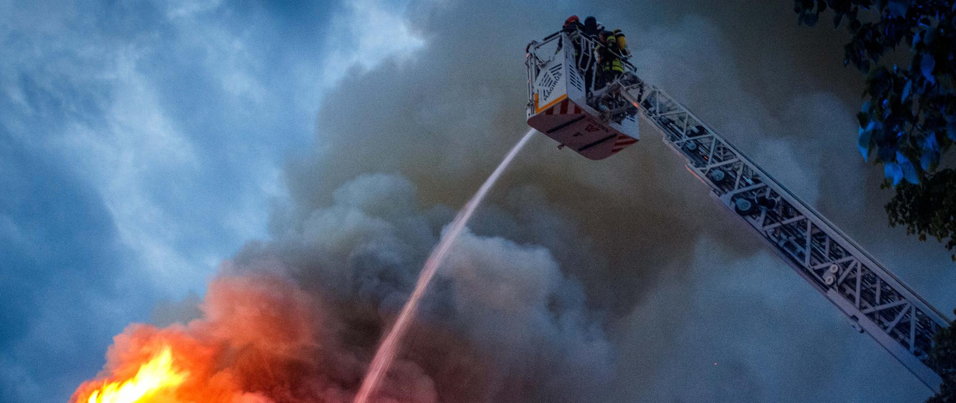 Zdjęcia przedstawia płonący budynek oraz strażaków którzy podają wodą z drabiny pożarniczej