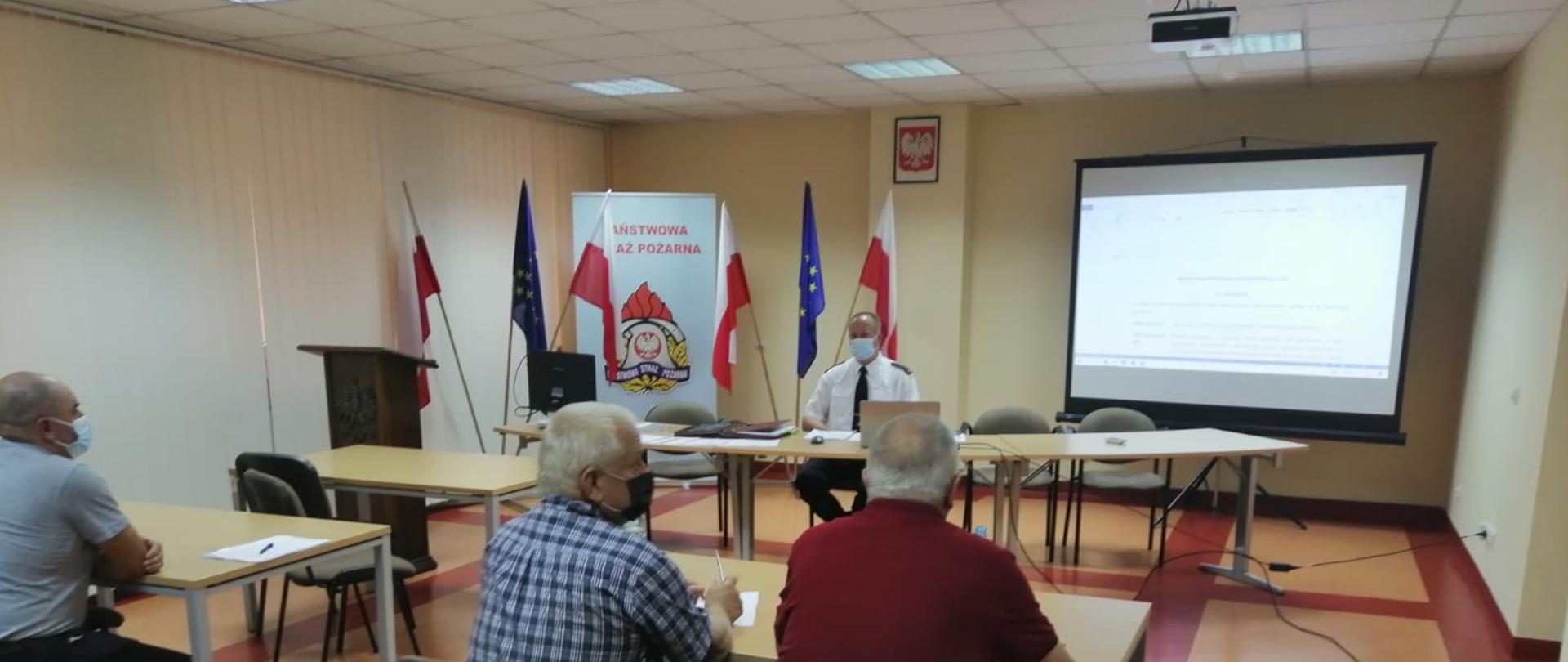 Zdjęcie przedstawia salę konferencyjną KP PSP w Jędrzejowie. Na sali zgromadzeni są przedstawiciele jednostek ochotniczych straży pożarnych z terenu powiatu jędrzejowskiego. Przed ochotnikami przy stole zasiada komendant powiatowy PSP w Jędrzejowie st. bryg. Paweł Jakubowski. Wszyscy mężczyźni mają założone maseczki ochronne zakrywające usta i nos.