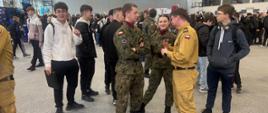 Na zdjęciu widoczna jest scena z wydarzenia odbywającego się w hali sportowej, w której uczestniczy liczna grupa młodzieży oraz przedstawiciele służb mundurowych. Na pierwszym planie widać rozmowę pomiędzy strażakiem w piaskowym mundurze a dwoma osobami ubranymi w mundury wojskowe. Rozmawiający stoją obok stanowiska prezentacyjnego, na którym znajdują się specjalistyczne narzędzia ratownicze, takie jak hydrauliczne rozpieracze i nożyce.
Wokół nich gromadzi się młodzież szkolna, która z zainteresowaniem obserwuje prezentacje i rozmawia z przedstawicielami służb. W tle widoczne są liczne stoiska informacyjne oraz inni uczestnicy wydarzenia związane z promocją służb.