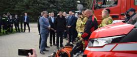 Grupa kilku osób stoi obok strażaków ubranych w mundury specjalne. Z prawej strony ustawione są samochody pożarnicze.