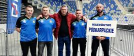 Zdjęcie przedstawia czterech zawodników drużyny województwa podkarpackiego w asyście kibica. W tle widoczna jest trybuna hali sportowej. Jeden z zawodników trzyma tabliczkę z napisem województwo podkarpackie.