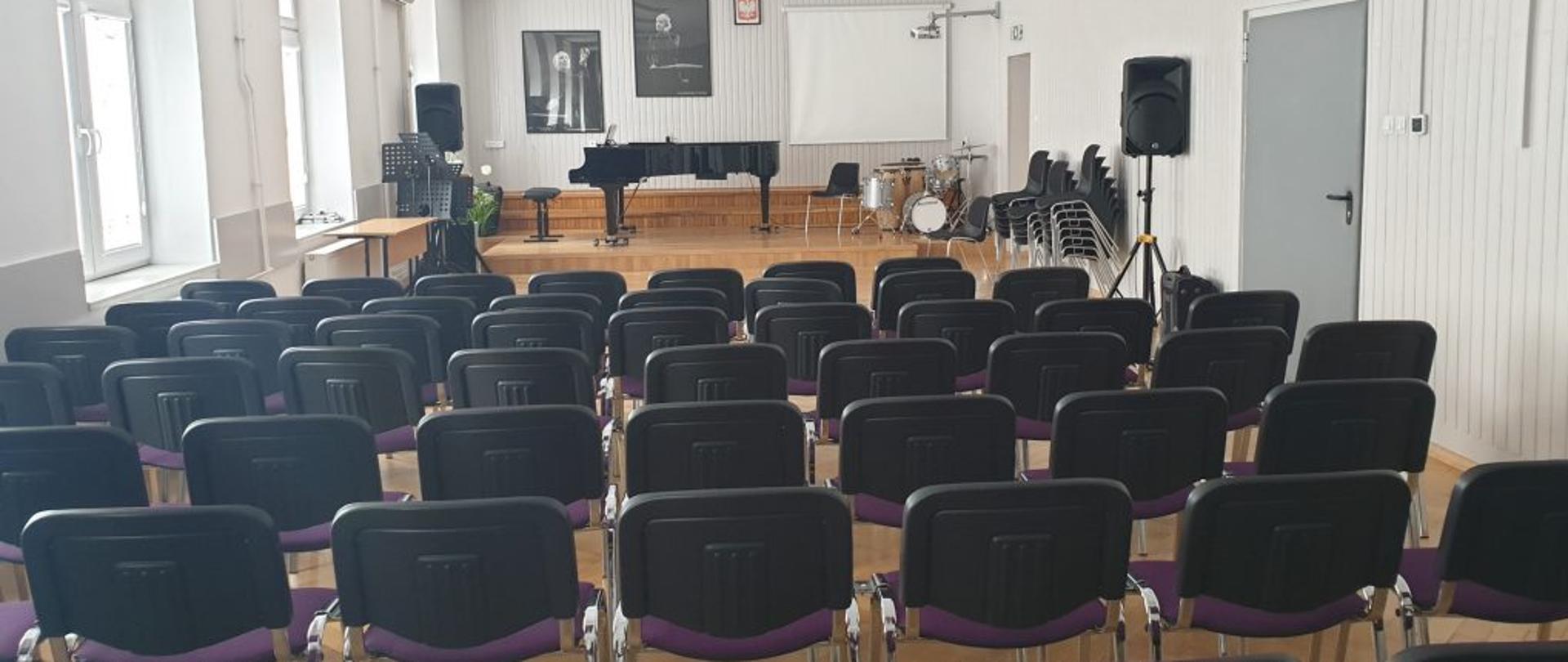 Na zdjęciu znajduje się scena w auli PSM, na której znajduje się fortepian, perkusja, pulpity i krzesła, na ścianie wiszą fotografie z Witoldem Lutosławskim oraz tablica interaktywna, na widowni sześć rzędów krzeseł