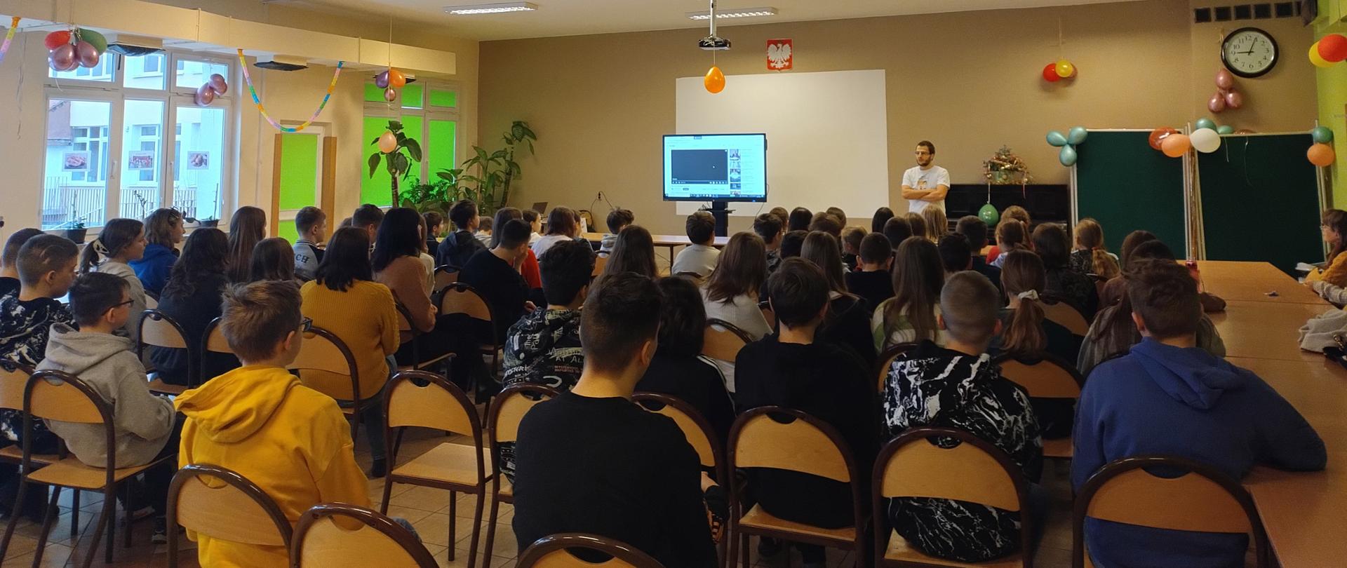 Dzieci siedzą w dużej sali na krzesłach naprzeciw interaktywnej tablicy. Obok niej stoi edukator Wód Polskich w białej koszulce z napisem Aktywni Błękitni. 