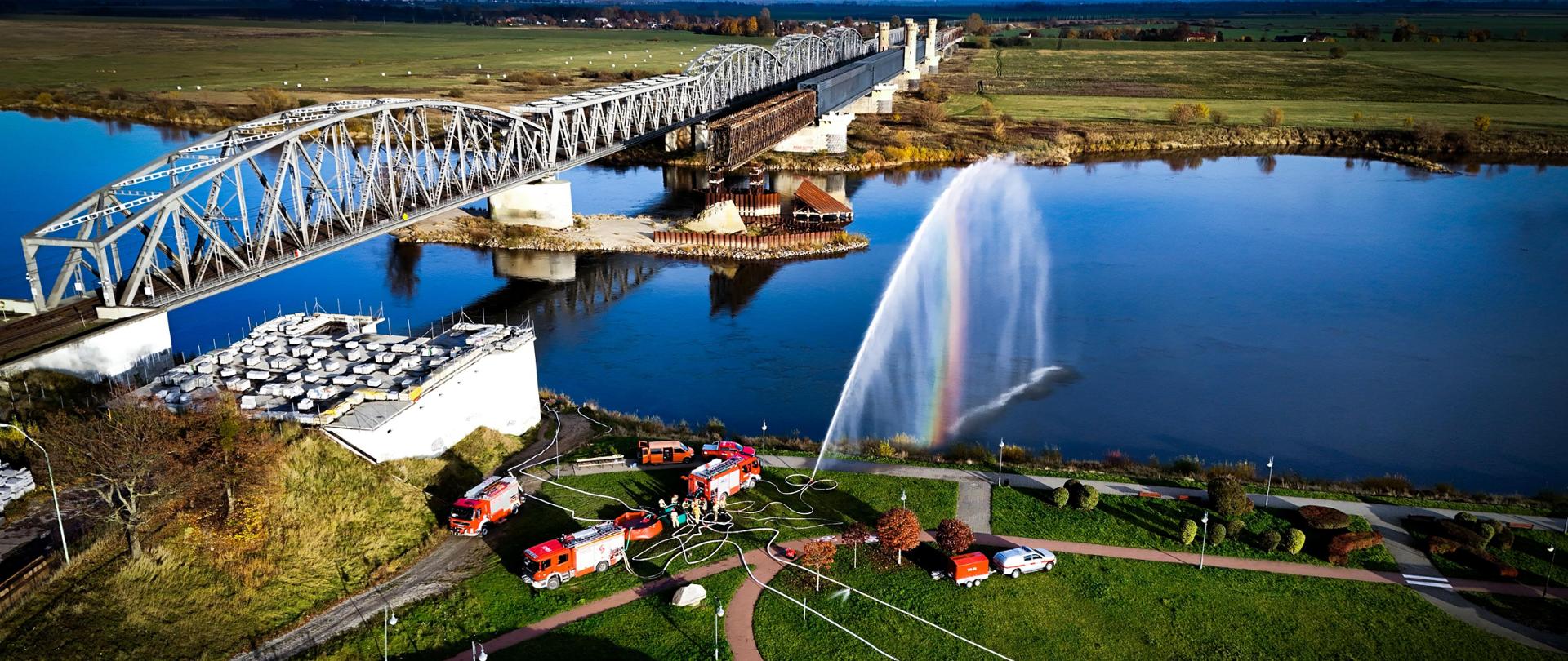 Ćwiczenia z przetłaczania wody, na zdjęciu 5 zastępów na bulwarze przy moście nad rzeką. Z działka leje się woda do rzeki - widok z drona.
