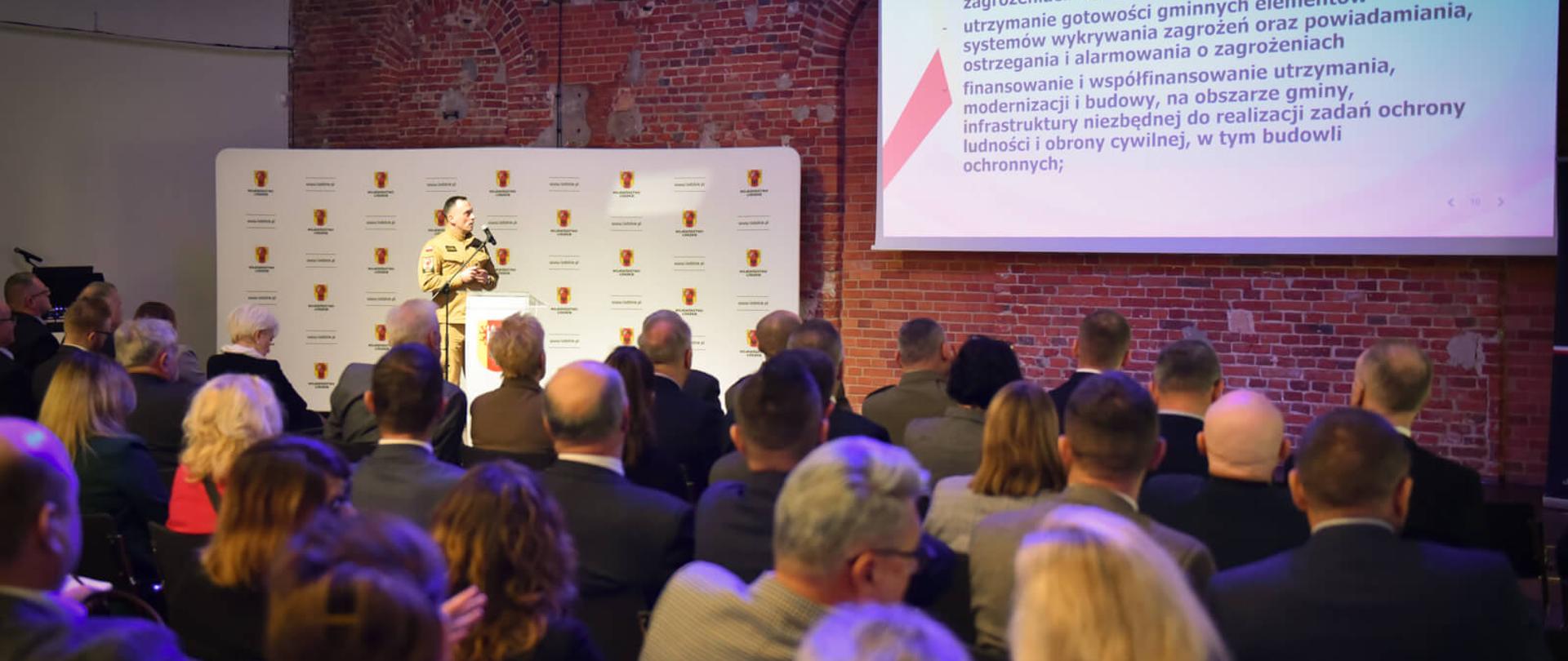 w dolnej części zdjęcia widać samorządowców siedzących w na krzesłach , w głębi po lewej, na tle ścianki, stoi starszy brygadier Mariusz Wojcieszko po prawej na ekranie rzutnika wyświetlany jest slajd 