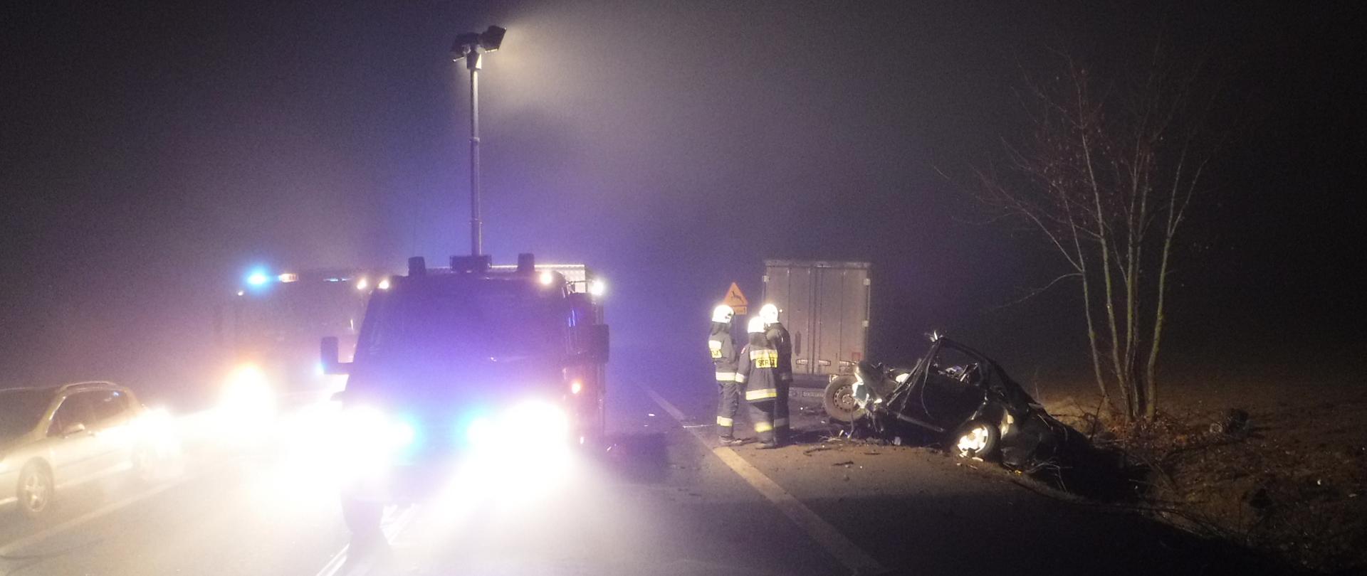 Na zdjęciu widać oświetlone miejsce wypadku (jest mgła), samochody strażackie oraz strażaków.