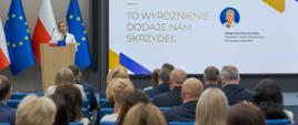 Duża sala konferencyjna , na której przemawia kobieta w białej marynarce. W tle baner Gwiazda Jakości.