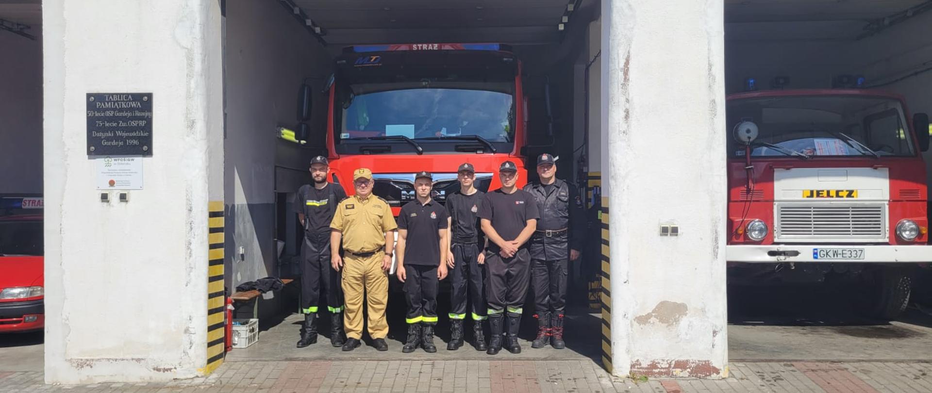 Strażacy Państwowej Straży Pożarnej oraz ochotniczych straży pożarnych stoją w bramie wyjazdowej przed samochodem gaśniczym obok otwarte są inne bramy a w nich stoją samochody gaśnicze. 