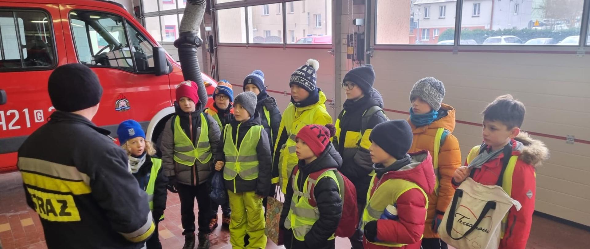 W garażu Jednostki Ratowniczo-Gaśniczej w Człuchowie na tle pojazdu pożarniczego stoi grupa kolonijna dzieci ze strażakiem, który opowiada im o służbie strażackiej.