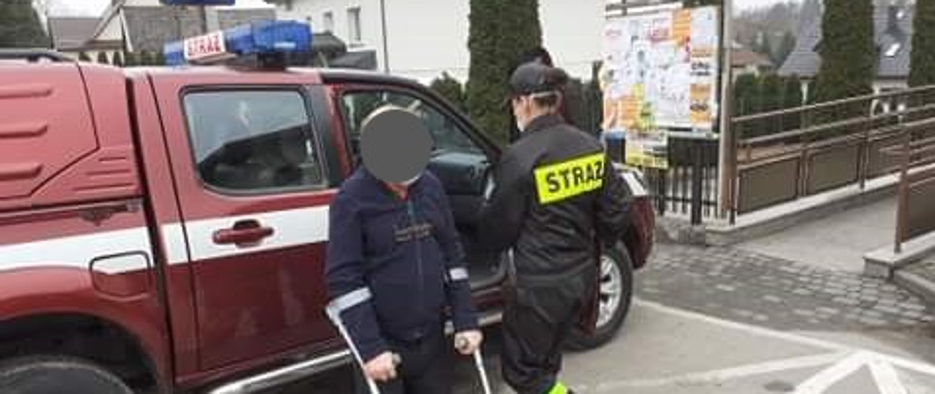 Na zdjęciu widzimy samochód strażacki przed którym stoi osoba dowieziona na szczepienie oraz odwrócony tyłem strażak. W tle widoczne zabudowania oraz tablica ogłoszeń.