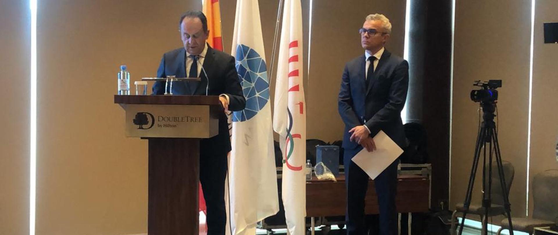 Na zdjęciu znajduje się dwóch elegancko ubranych mężczyzn. Mężczyźni stoją na podeście sceny, z czego jeden z nich jest w trakcie wystąpienia (stoi za mównicą). W tle znajdują się trzy flagi - Macedonii Północnej w barwach żółto-czerwonych oraz białe flagi komitetów olimpijskich z ich logotypami. 