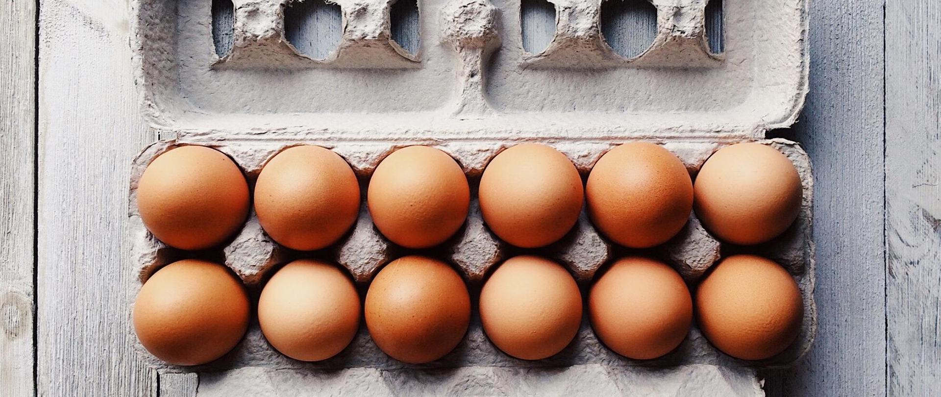 Na zdjęciu znajduje się otwarta tekturowa wytłoczka z dwunastoma świeżymi jajami. Wytłoczka jest na drewnianych deskach. Widok na wytłoczkę i jaja z góry.