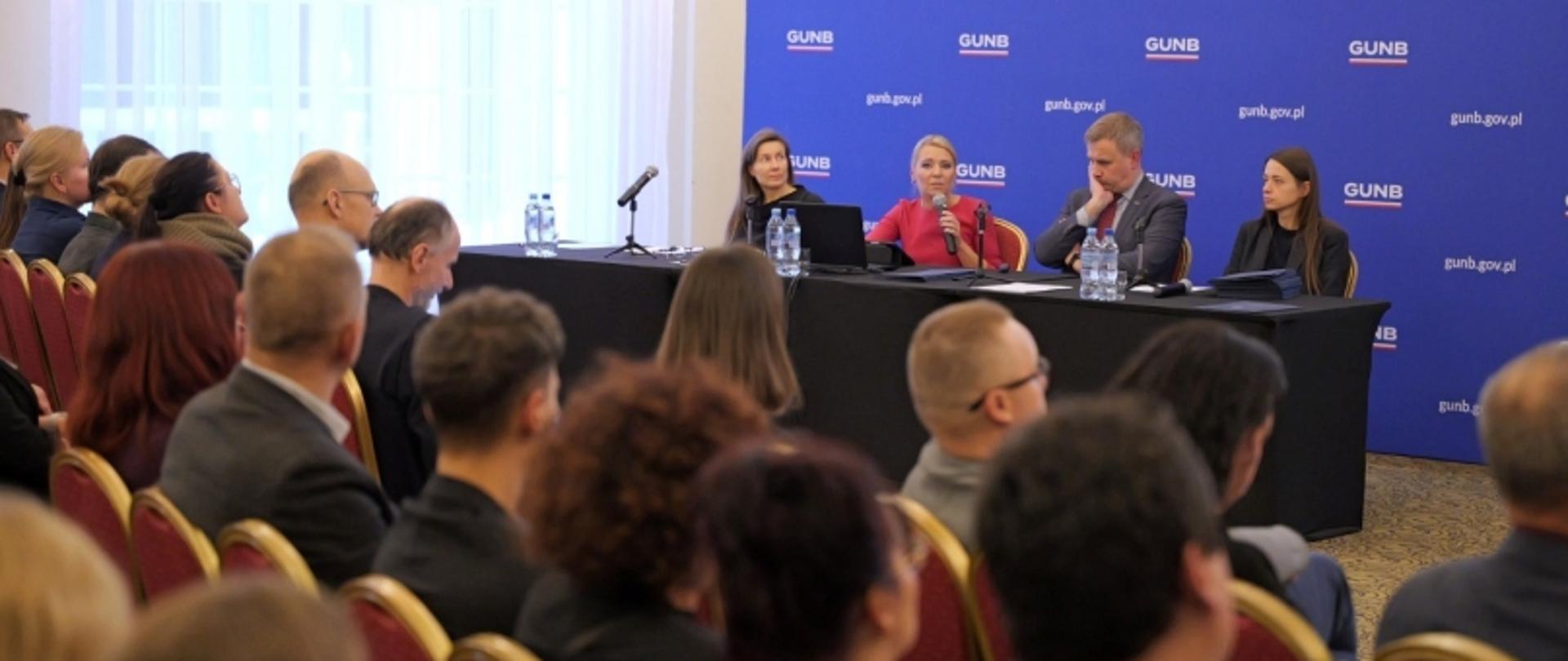 Cztery osoby przy stole konferencyjnym z mikrofonami, przed nimi publiczność na czerwonych krzesłach; w tle baner GUNB z adresem gunb.gov.pl.