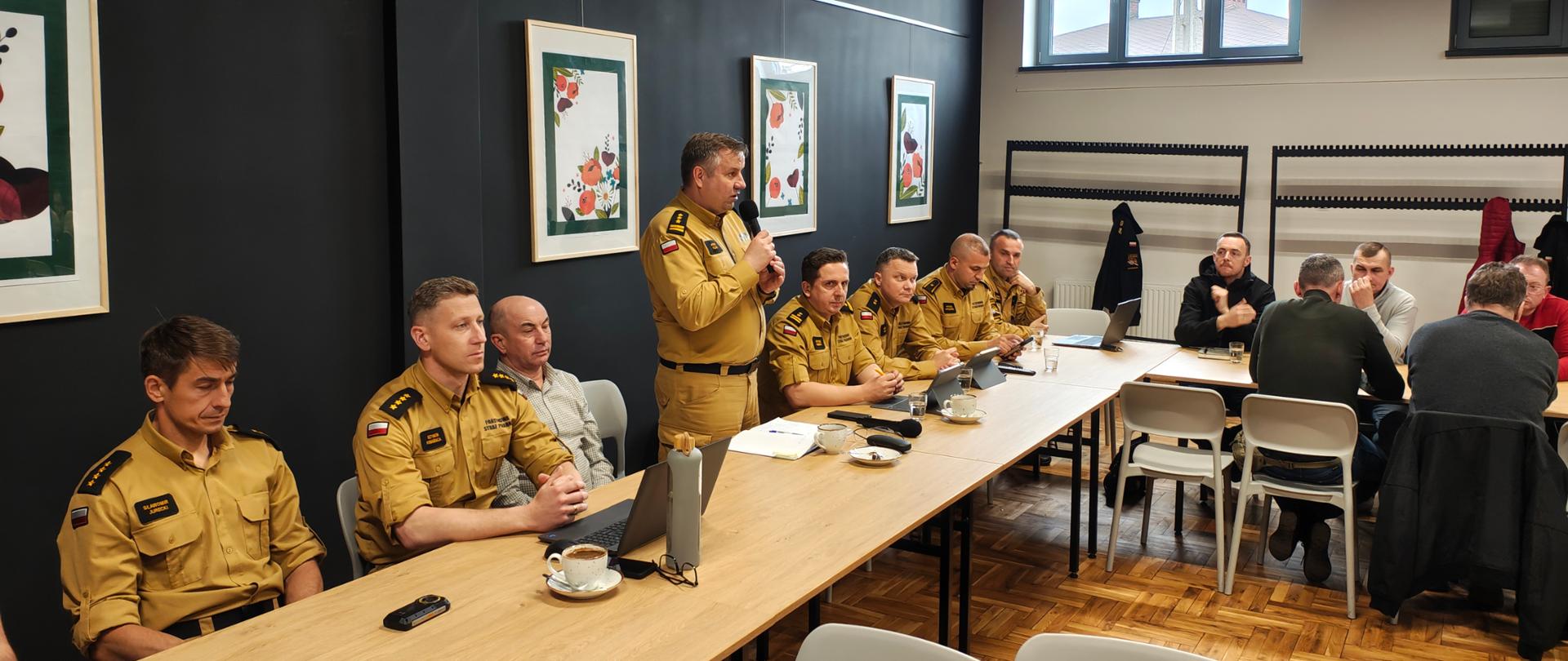 Spotkanie w sali konferencyjnej: kilku umundurowanych strażaków siedzi przy długim stole, jeden z nich przemawia do mikrofonu. Na stole znajdują się laptopy, dokumenty i filiżanki z kawą. W tle widać innych uczestników spotkania, okno oraz ściany ozdobione oprawionymi grafikami.