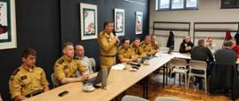 Spotkanie w sali konferencyjnej: kilku umundurowanych strażaków siedzi przy długim stole, jeden z nich przemawia do mikrofonu. Na stole znajdują się laptopy, dokumenty i filiżanki z kawą. W tle widać innych uczestników spotkania, okno oraz ściany ozdobione oprawionymi grafikami.