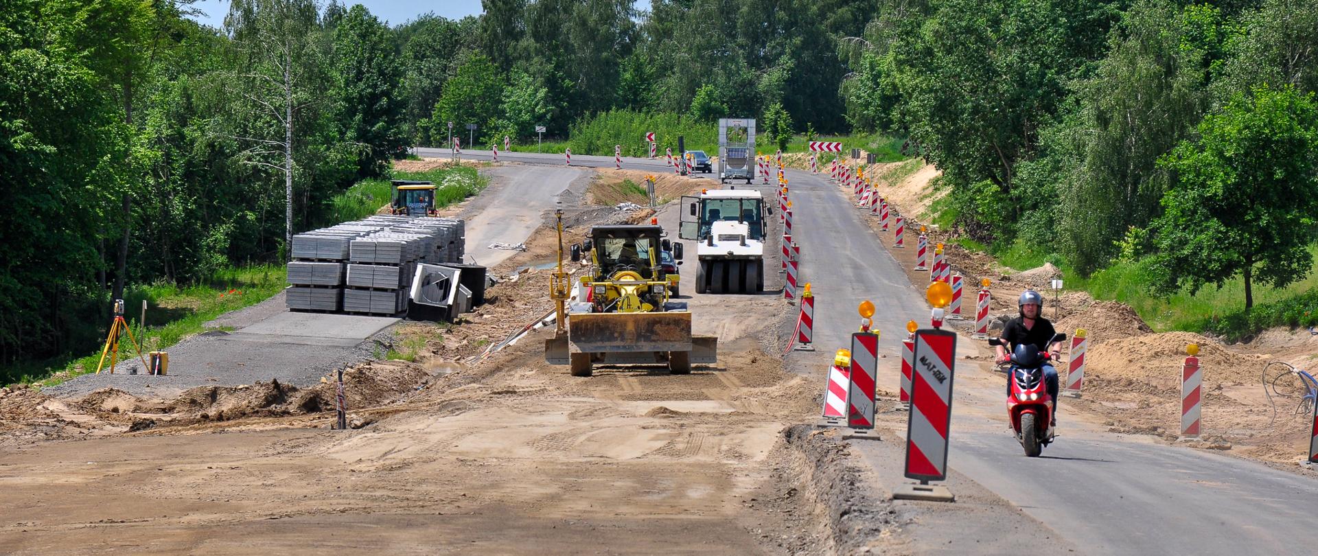 Zdjęcie przedstawia fragment drogi krajowej DK74 w miejscowości Gorajec-Szczebrzeszyn. Na pierwszym planie widoczna jest jezdnia z oznakowaniem poziomym, w tym linie rozdzielające pasy ruchu. Po obu stronach drogi znajdują się pobocza z trawą oraz słupki drogowe. W tle widoczne są drzewa i krzewy, a także elementy infrastruktury drogowej, takie jak znaki drogowe. Kolorystyka naturalna – zielone rośliny, szara nawierzchnia asfaltowa, niebo w odcieniach jasnych (prawdopodobnie pochmurne lub częściowo słoneczne).