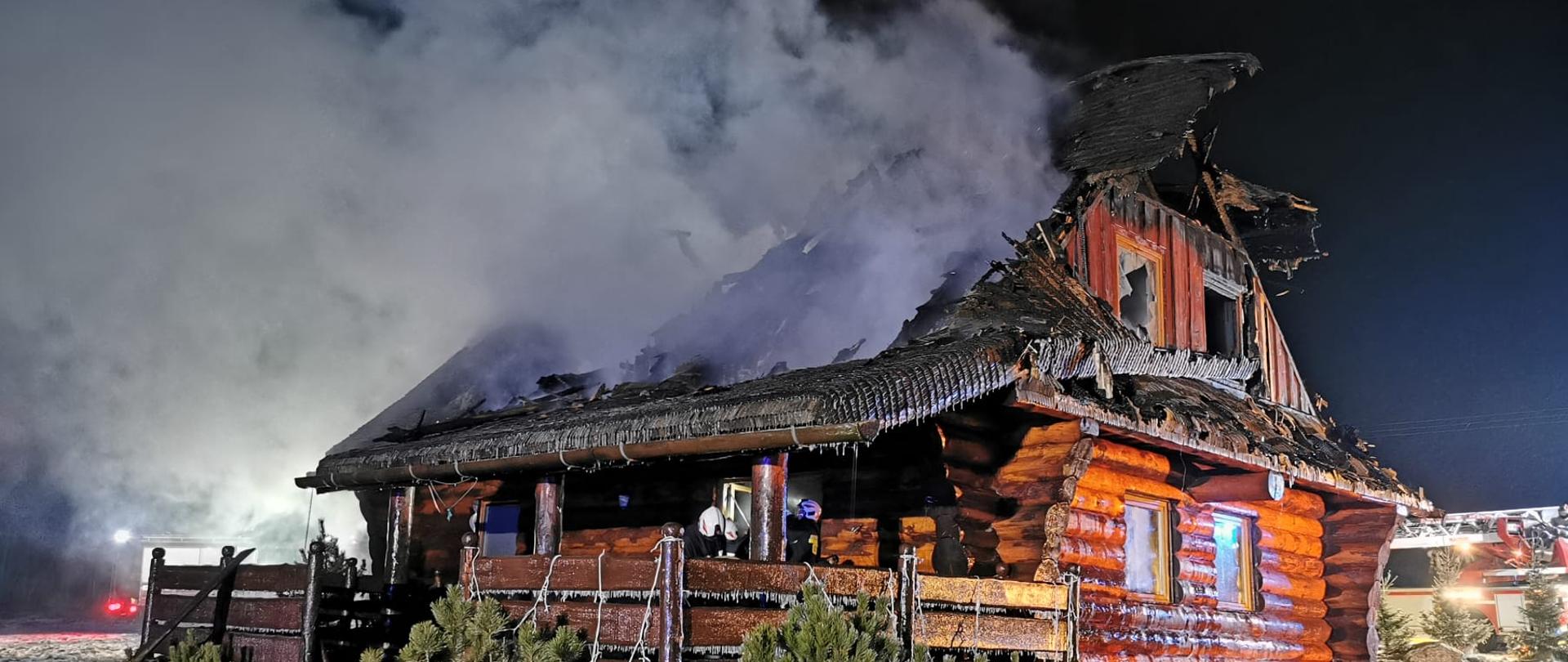 Pora nocna. Widok na zniszczony wskutek pożaru drewniany budynek jednorodzinny. Ze spalonego dachu wydobywają się kłęby dymu.