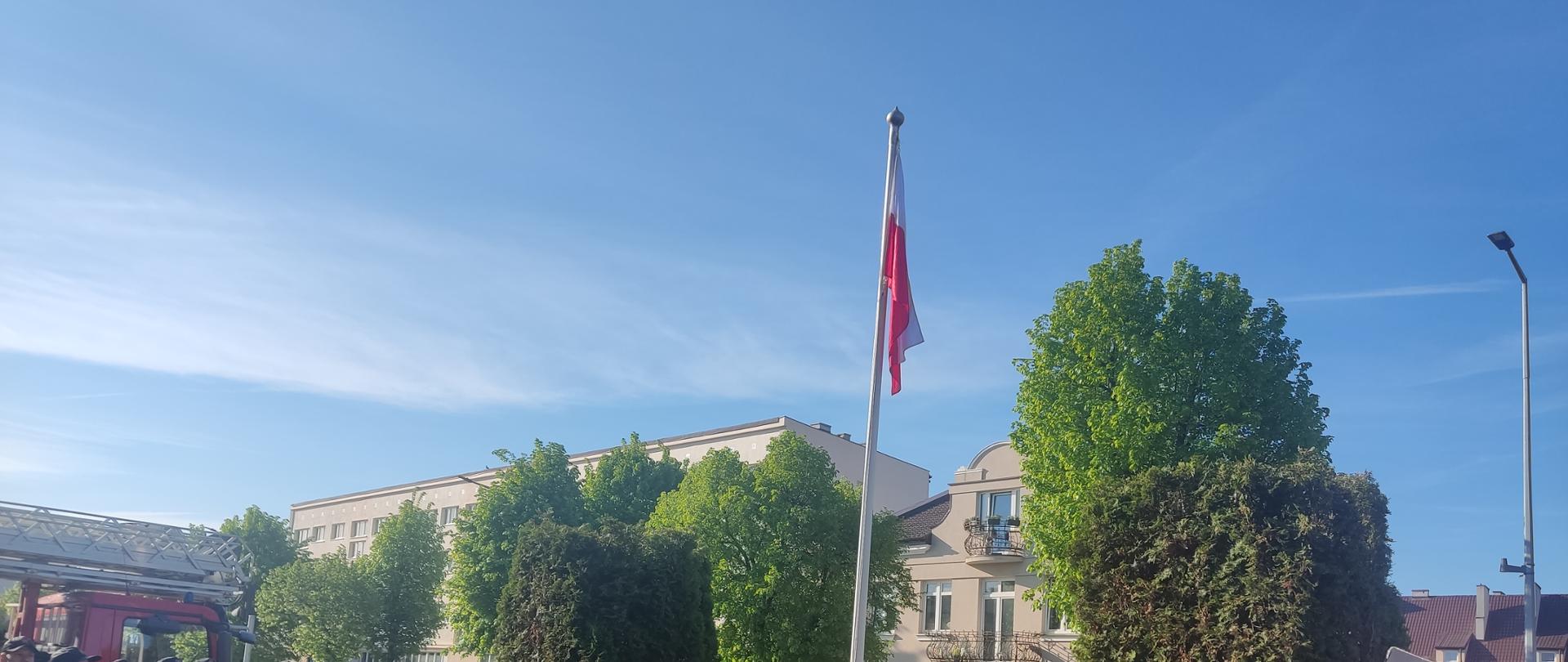 Zdjęcie przedstawia strażaków podczas uroczystego apelu z okazji Dnia Flagi Rzeczypospolitej Polskiej i flagę państwową wciągniętą na maszt