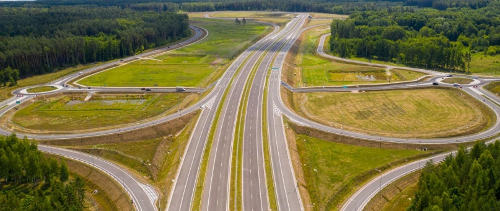 Aerial view na węzeł autostradowy z wieloma pasami i zakrętami, otoczony lasami i polami; kilka samochodów na drogach dojazdowych, główna trasa pusta.