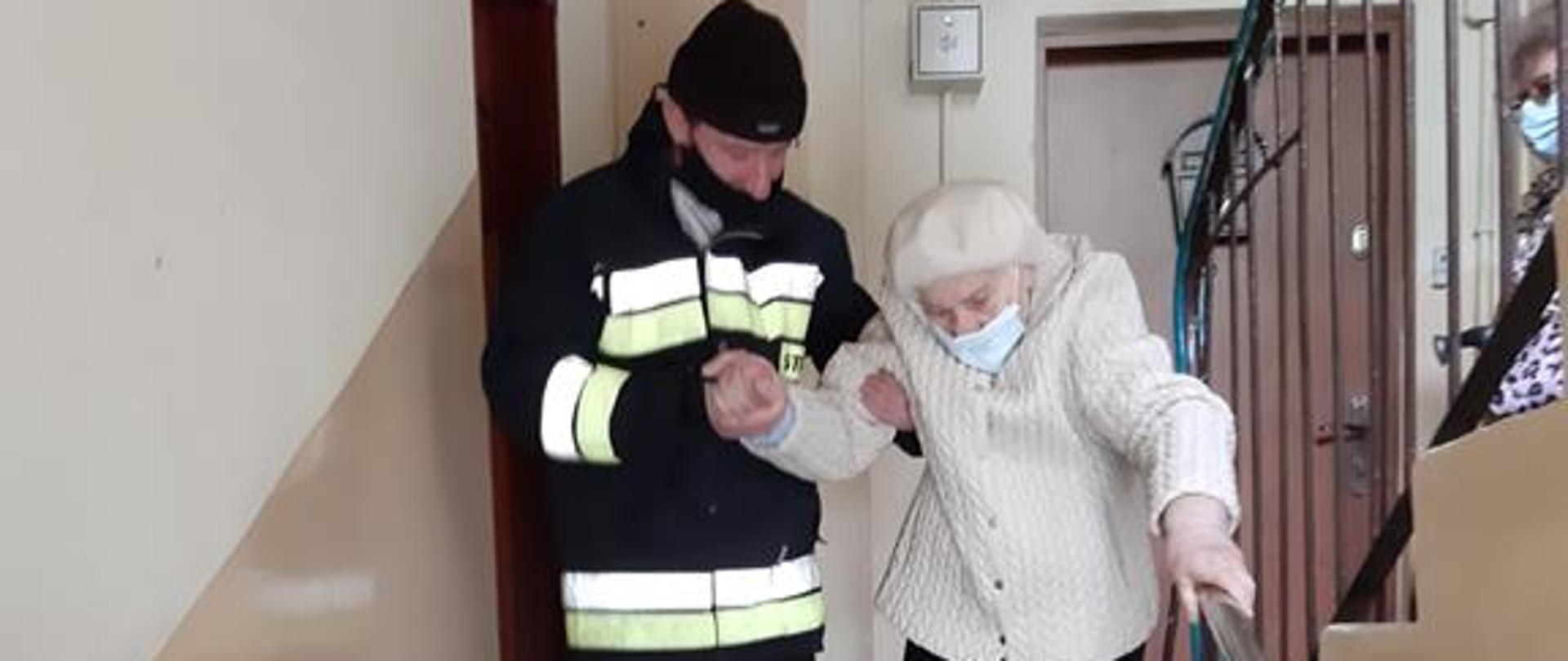 Strażak ratownik w ubraniu specjalnym pomaga starszej kobiecie zejść po klatce schodowej. Kobieta przytrzymuje się lewą ręką poręczy a drugą jest asekurowana przez strażaka