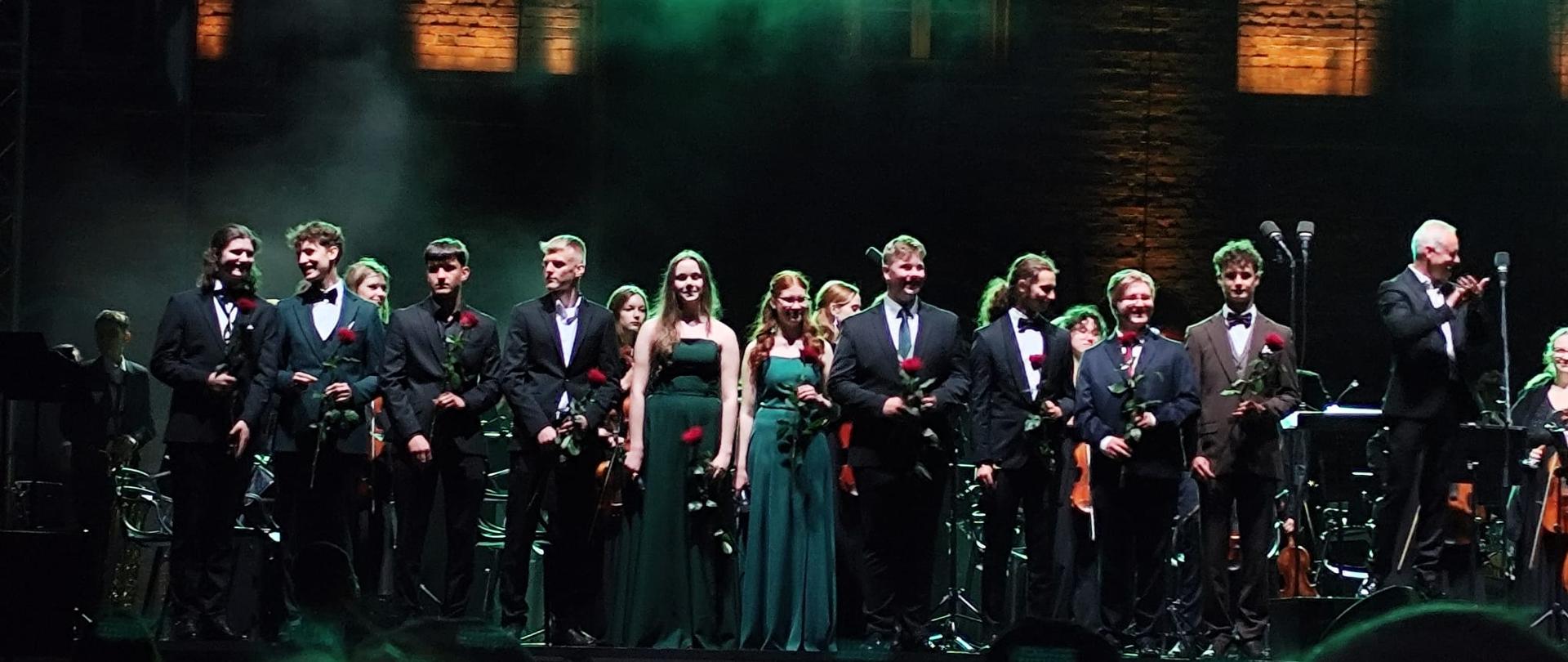 Na scenie podczas koncertu grupa młodych muzyków i wokalistów w eleganckich strojach, trzymających czerwone róże. W tle widoczna orkiestra, a scenę oświetla zielone światło.