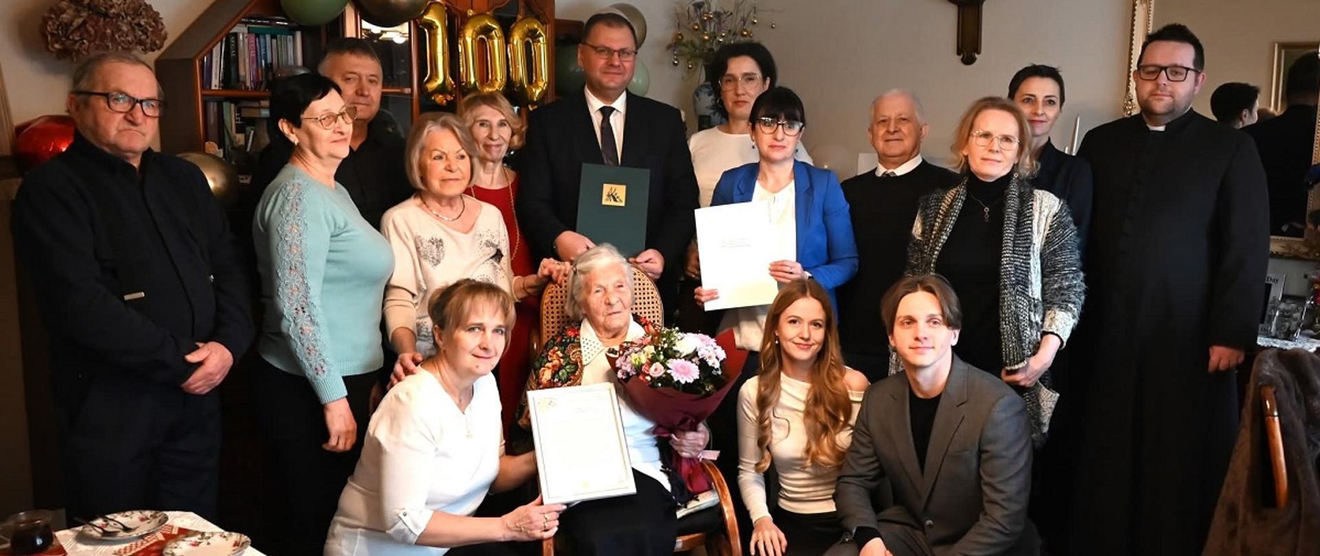 1 Kilkanaście osób zgromadzonych w salonie podczas uroczystości jubileuszowej. W centrum siedzi starsza kobieta trzymająca bukiet kwiatów oraz dokument, otoczona przez rodzinę i zaproszonych gości. Za zgromadzonymi widoczne są złote balony z liczbą „100”, symbolizujące setne urodziny. Część osób trzyma oficjalne pisma lub teczki, inni stoją lub klęczą obok jubilatki. W tle widać meble, regał z książkami i dekoracje.