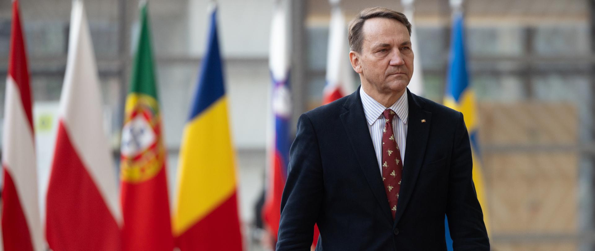 Mężczyzna w garniturze i krawacie idzie korytarzem budynku instytucji międzynarodowej; w tle stoją flagi kilku państw, w tym Polski, Portugalii, Rumunii i Szwecji.