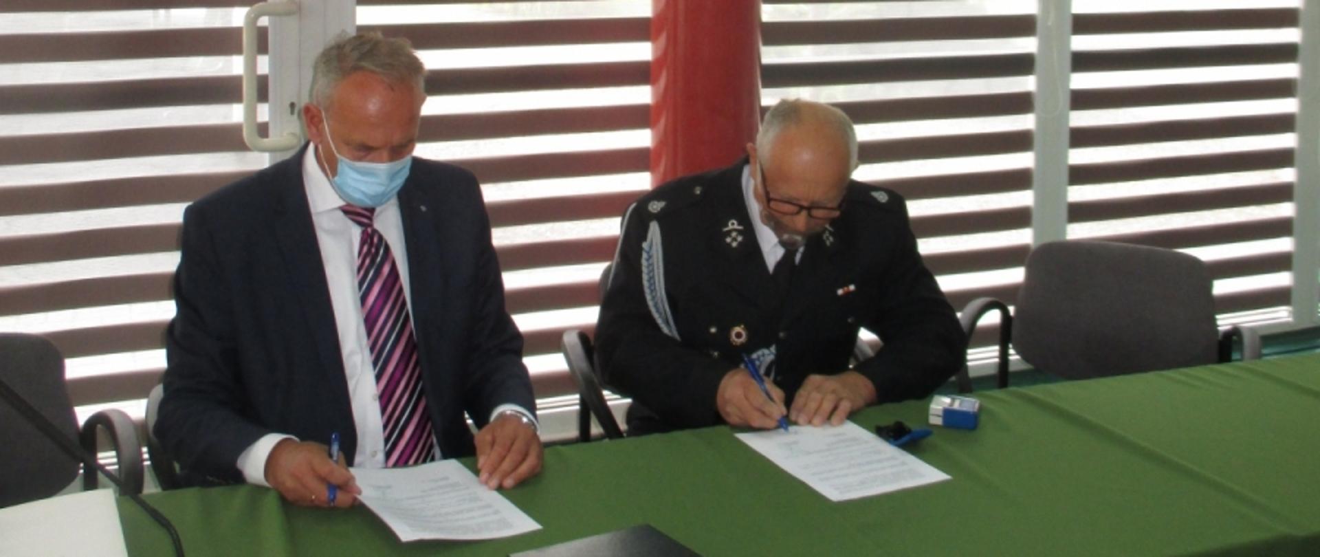 Za zdjęciu moment podpisania umowy. Przy stole prezydialnym po lewej stronie siedzi Pan Andrzej Skiba Prezes Zarządu Wojewódzkiego Funduszu Ochrony Środowiska i Gospodarki Wodne w Rzeszowie trzymając w prawej ręce długopis i umowę. Po prawej stronie Prezes Ochotniczej Straży Pożarnej w Gliniku Średnim siedząc przy stole podpisuje umowę. Obaj Panowie w maseczkach ochronnych. Na stole w kolorze zielonym widać od lewej strony: białą teczkę na dokumenty, mikrofon, czarą teczkę na dokumenty, dwie umowy oraz pieczątkę.
