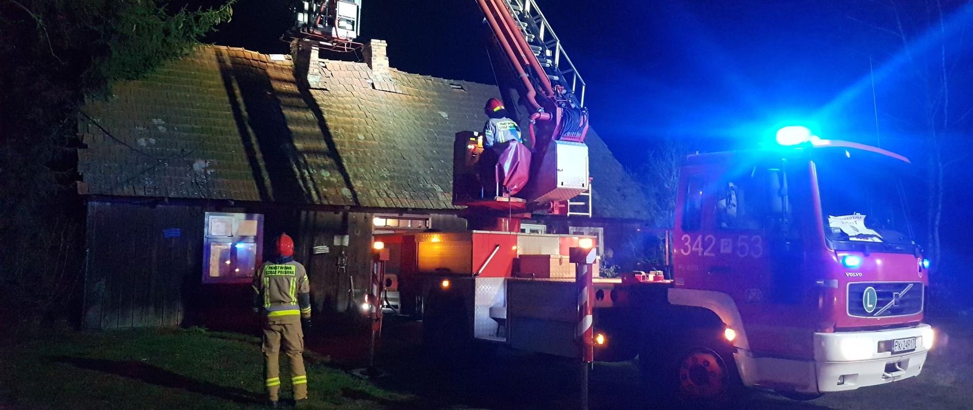 Zdjęcie wykonane w porze nocnej. Widać pojazdy straży pożarnej w kolorze czerwonym, z oznaczeniami. Na drugim planie zabudowania. Podłoże trawiaste. Występują źródła światła sztucznego z pojazdów straży pożarnej.