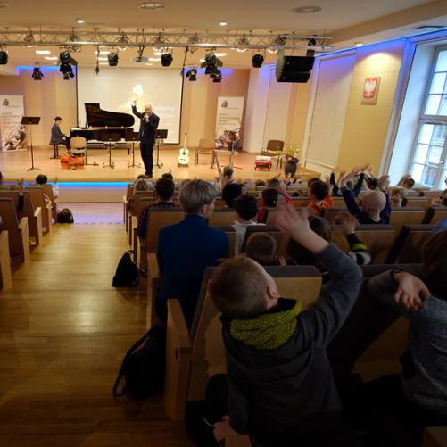 zdjęcie przedstawia prowadzącego koncert na scenie sali koncertowej podczas zabawy z uczestnikami koncertu siedzącymi na widowni, przy fortepianie akompaniator, na scenie instrumenty oraz banery szkoły i w tle ekran multimedialny z plakatem koncertu 