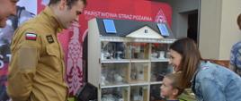 Dzień otwarty w rawickiej komendzie. Stanowisko z mobilnym symulatorem zagrożeń pożarowych, który ustawiony jest na tle ścianku promującej KP PSP w Rawiczu. Przed symulatorem stoi mama z kilkuletnim synem. Słuchają wiadomości na temat zagrożeń, przekazywanych przez strażaka ubranego w mundur koloru piaskowego. Pani usmiecha się.