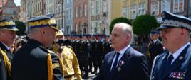 Strażak w mundurze galowym gratuluje odznaczenia mężczyźnie w garniturze obok stoi przedstawiciel Policji niedaleko stoją wyróżnieni funkcjonariusze Państwowej Straży Pożarnej.