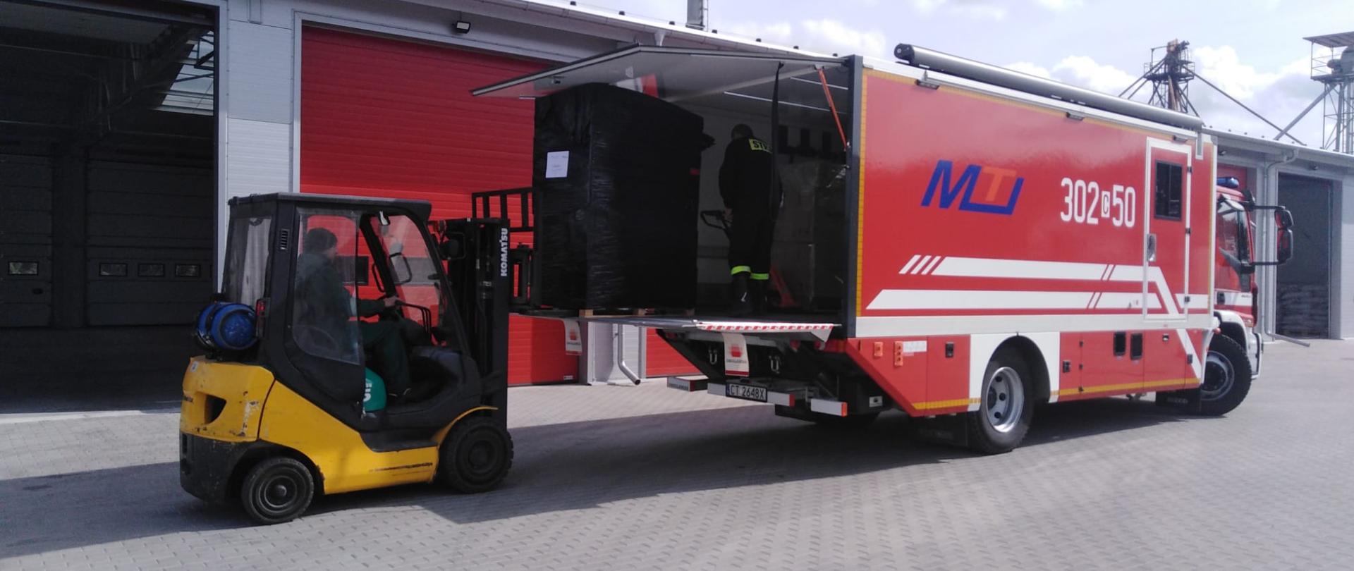 Z lewej strony widać żółty wózek widłowy który ładuje na pakę samochodu specjalnego marki Iveco paletę. Widać prawy bok pojazdu Iveco. Obydwa pojazdy stoją na kostce, w tle niski budynek magazynowy z jasną siwą elewacją i czerwonymi bramami. 