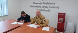 Przy stole siedzi komendant powiatowy Państwowej Straży Pożarnej w Działdowie oraz jeden ze strażaków-ochotników. Przeglądają kartki ze spisaną umową na pełnienie funkcji kierowcy w Ochotniczej Straży Pożarnej 