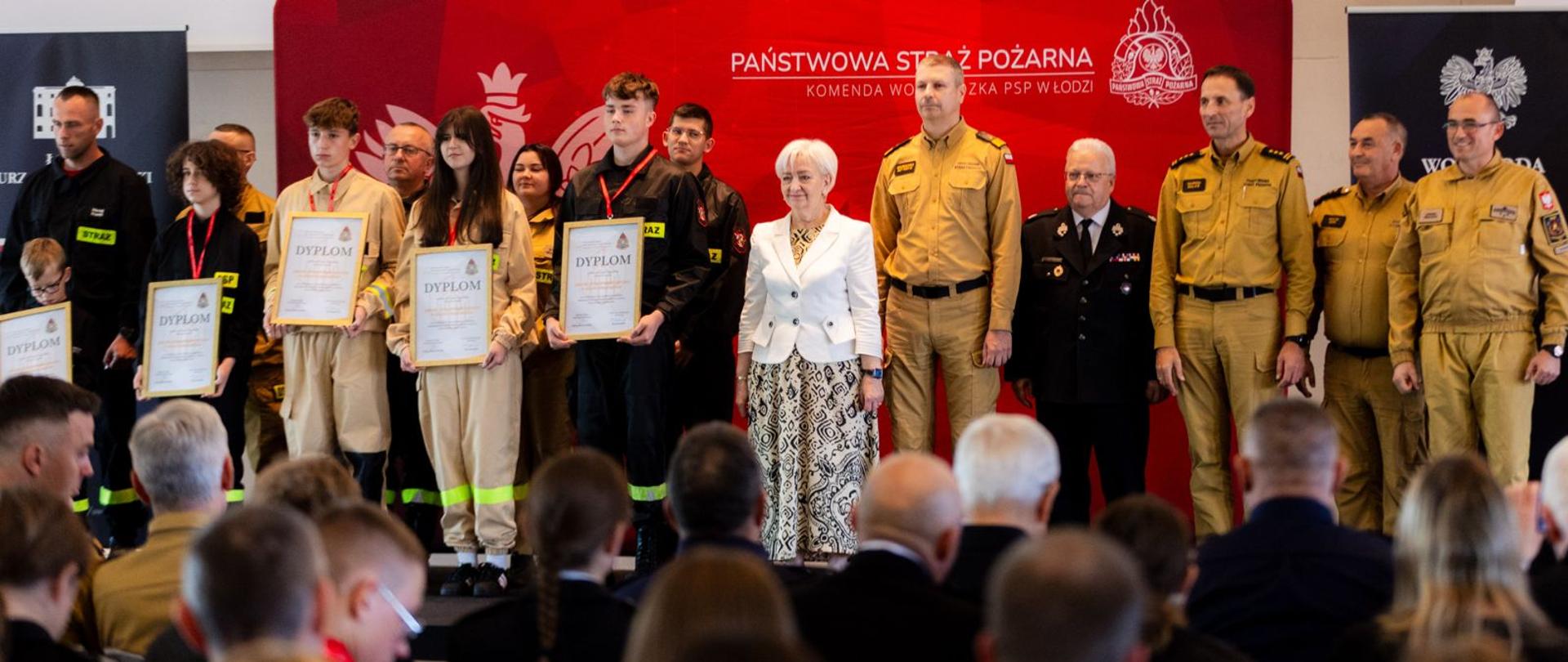 Ujęcie z perspektywy widowni na grupę nagrodzonych osób i oficjeli stojących na scenie. Młodzież trzyma oprawione w ramy dyplomy.